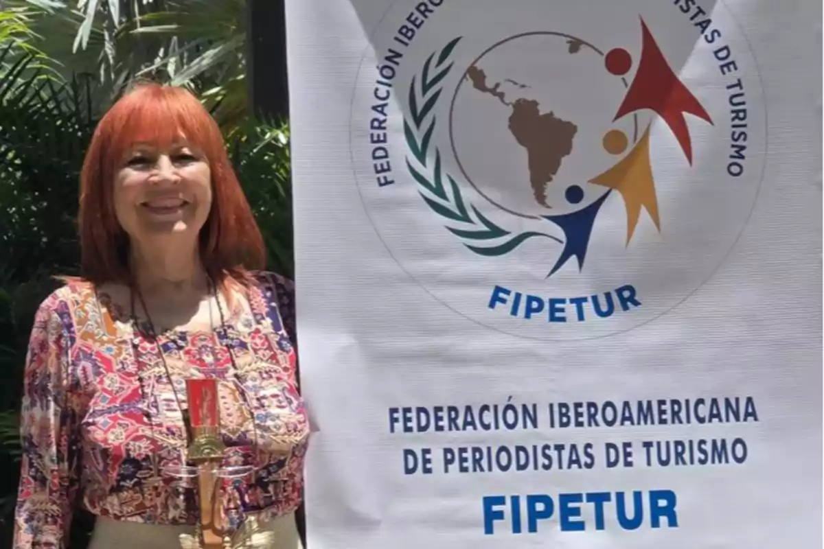 Una persona sonriente está de pie junto a un cartel de la Federación Iberoamericana de Periodistas de Turismo (FIPETUR) en un entorno al aire libre con vegetación.