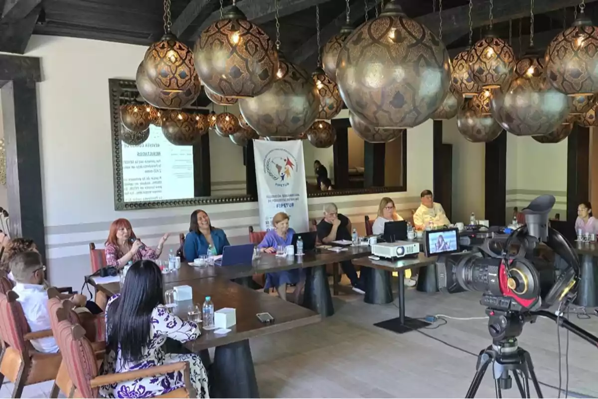 Un grupo de personas está reunido en una sala de conferencias decorada con lámparas colgantes, hay una cámara de video en primer plano y un proyector sobre la mesa. Un grupo de personas está reunido en una sala de conferencias decorada con lámparas colgantes, hay una cámara de video en primer plano y un proyector sobre la mesa.