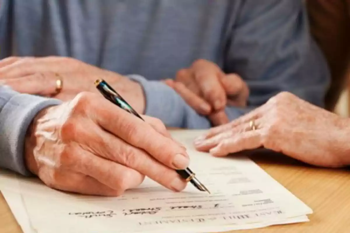 Persona mayor firmando un documento con una pluma mientras otra persona le acompaña.