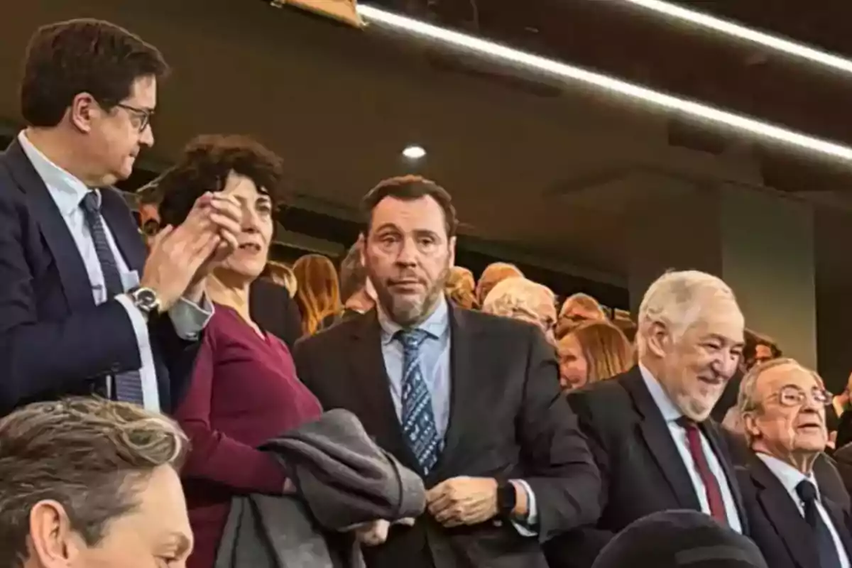 Un grupo de personas vestidas formalmente está de pie en lo que parece ser un evento o reunión, con una multitud de personas al fondo. Un grupo de personas vestidas formalmente está de pie en lo que parece ser un evento o reunión, con una multitud de personas al fondo.