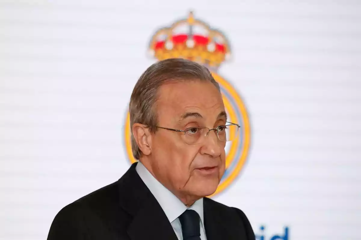 Un hombre con gafas y traje oscuro hablando frente a un fondo con un escudo.