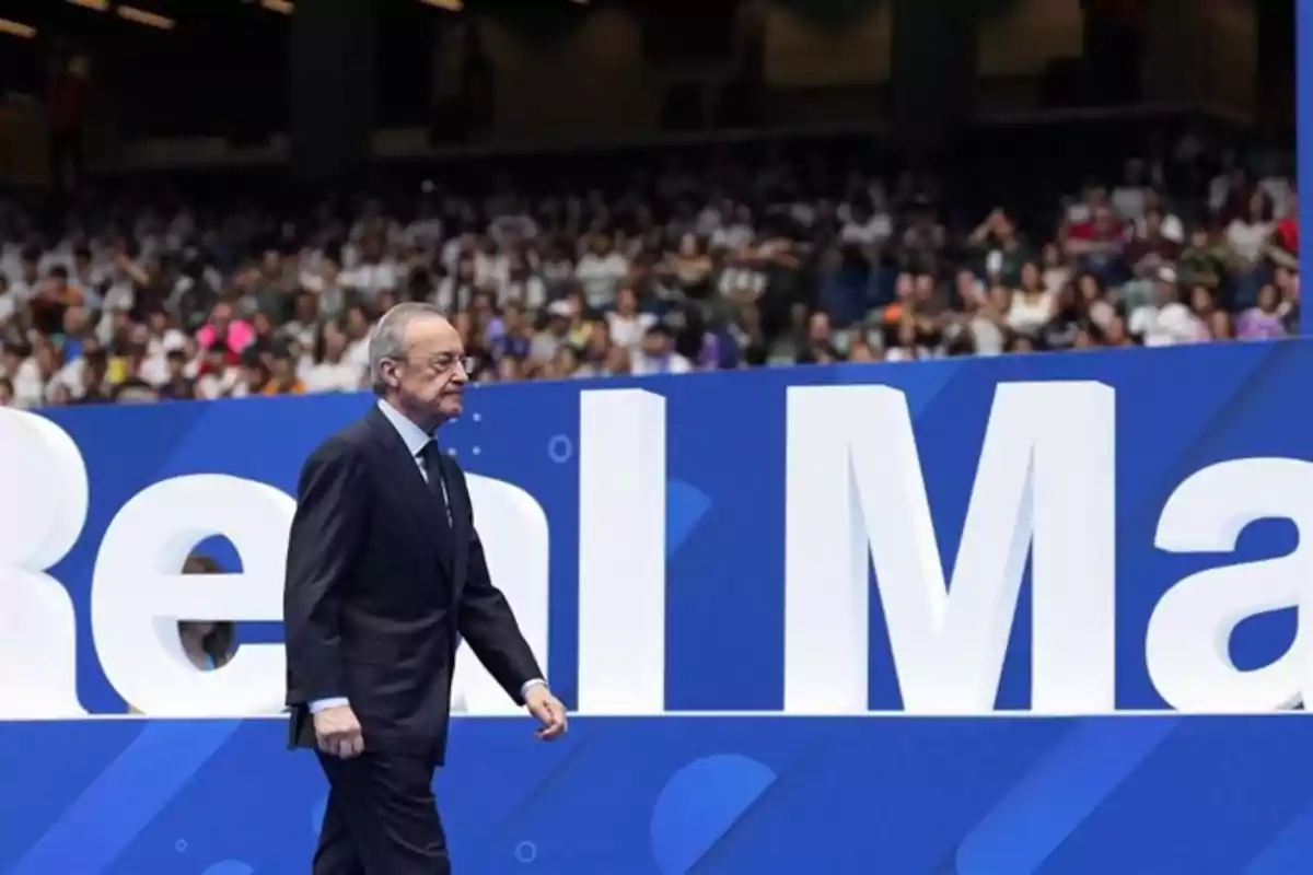 Un hombre caminando frente a un cartel azul con letras grandes y una multitud de personas al fondo.