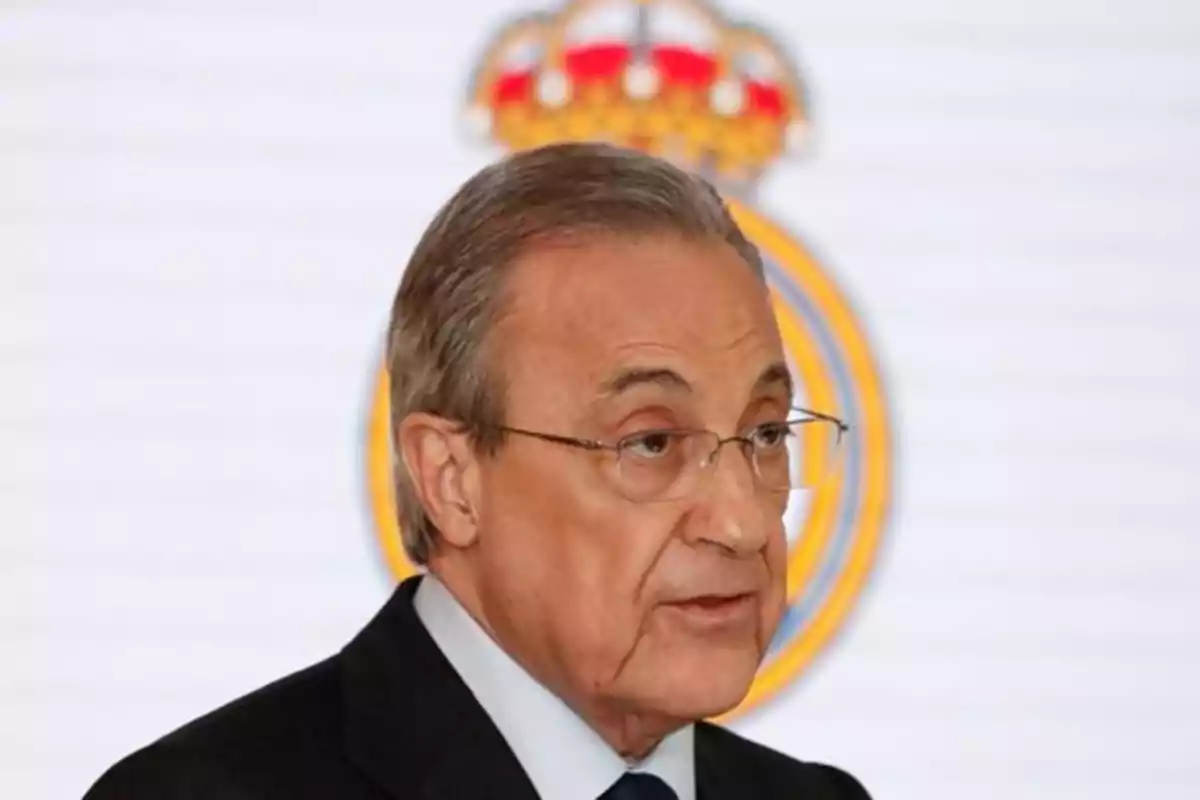 Un hombre con gafas y traje oscuro hablando frente a un fondo con un escudo.