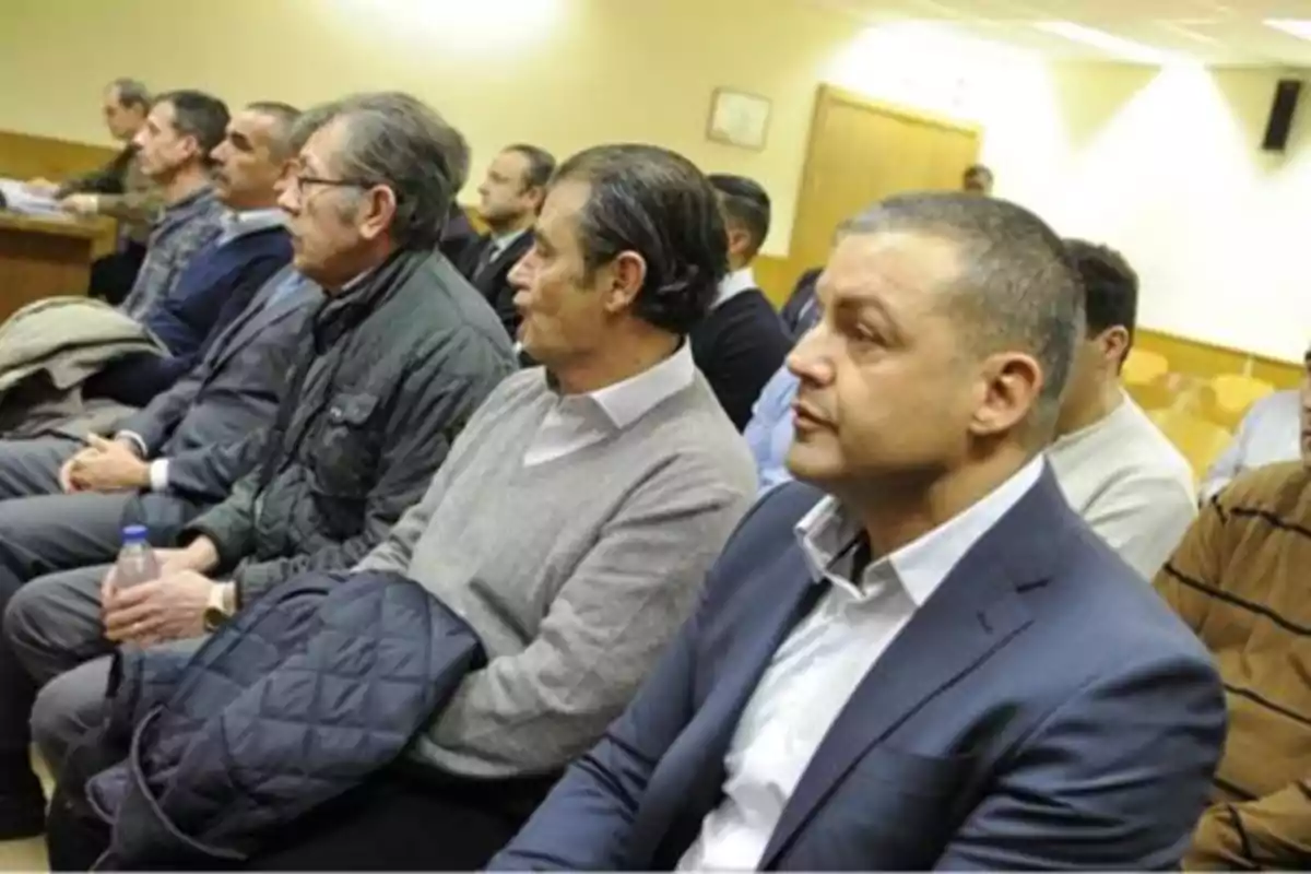 Un grupo de hombres sentados en una sala, algunos con chaquetas y suéteres, mirando hacia el frente.