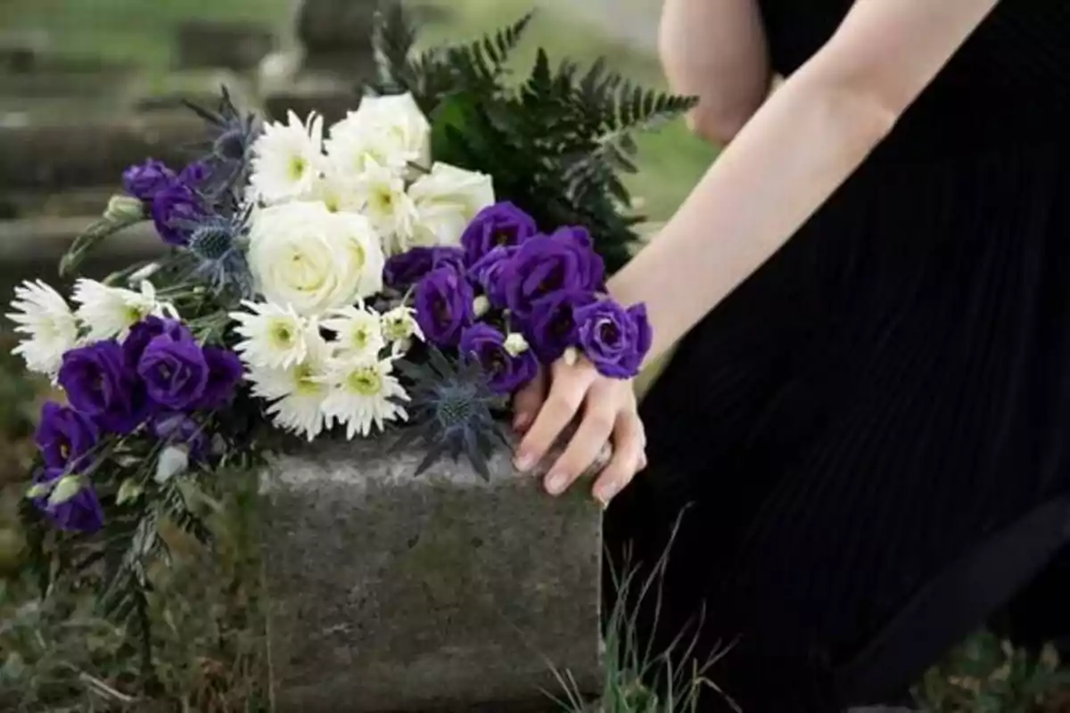 Una persona colocando un ramo de flores blancas y moradas sobre una lápida en un cementerio.