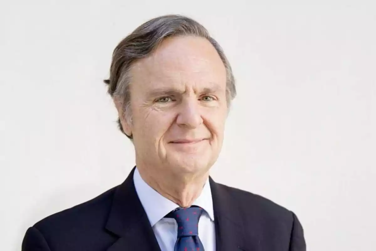 Hombre de mediana edad con traje oscuro y corbata azul sonriendo ligeramente frente a un fondo claro.