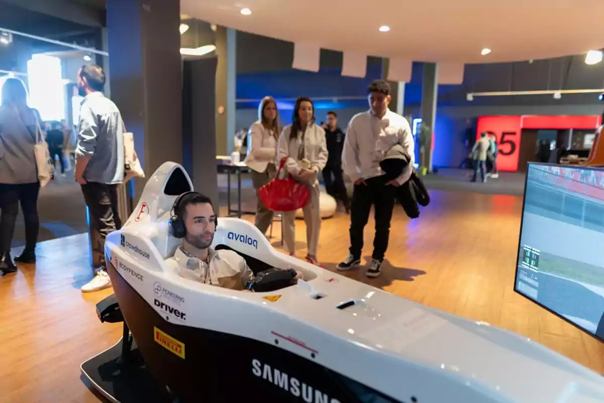 Un hombre está sentado en un simulador de carreras de autos mientras otras personas observan a su alrededor en un entorno interior iluminado.