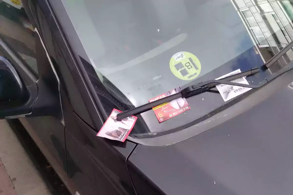 Un coche negro estacionado con varios folletos publicitarios sujetos bajo el limpiaparabrisas delantero. Un coche negro estacionado con varios folletos publicitarios sujetos bajo el limpiaparabrisas delantero.