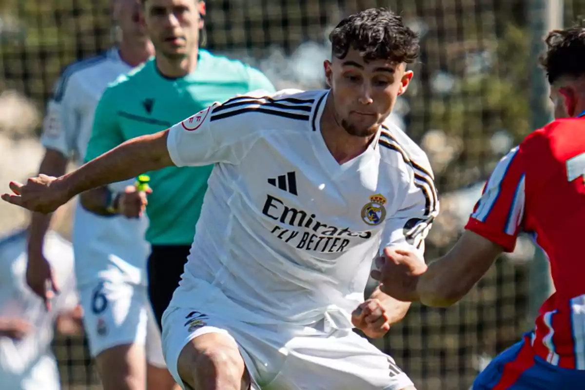 Jugador del Real Madrid con uniforme blanco disputa el balón contra un rival con uniforme rojo durante un partido de fútbol mientras un árbitro observa la jugada al fondo Jugador del Real Madrid con uniforme blanco disputa el balón contra un rival con uniforme rojo durante un partido de fútbol mientras un árbitro observa la jugada al fondo