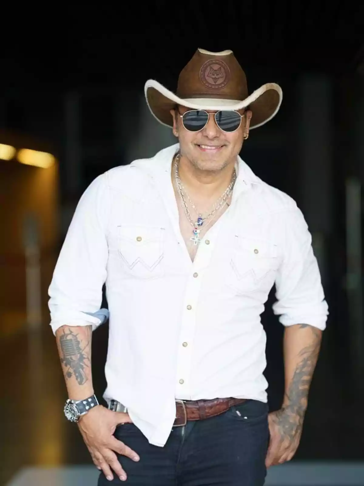 Un hombre con sombrero vaquero, gafas de sol y camisa blanca sonríe mientras posa con una mano en el bolsillo. Un hombre con sombrero vaquero, gafas de sol y camisa blanca sonríe mientras posa con una mano en el bolsillo.