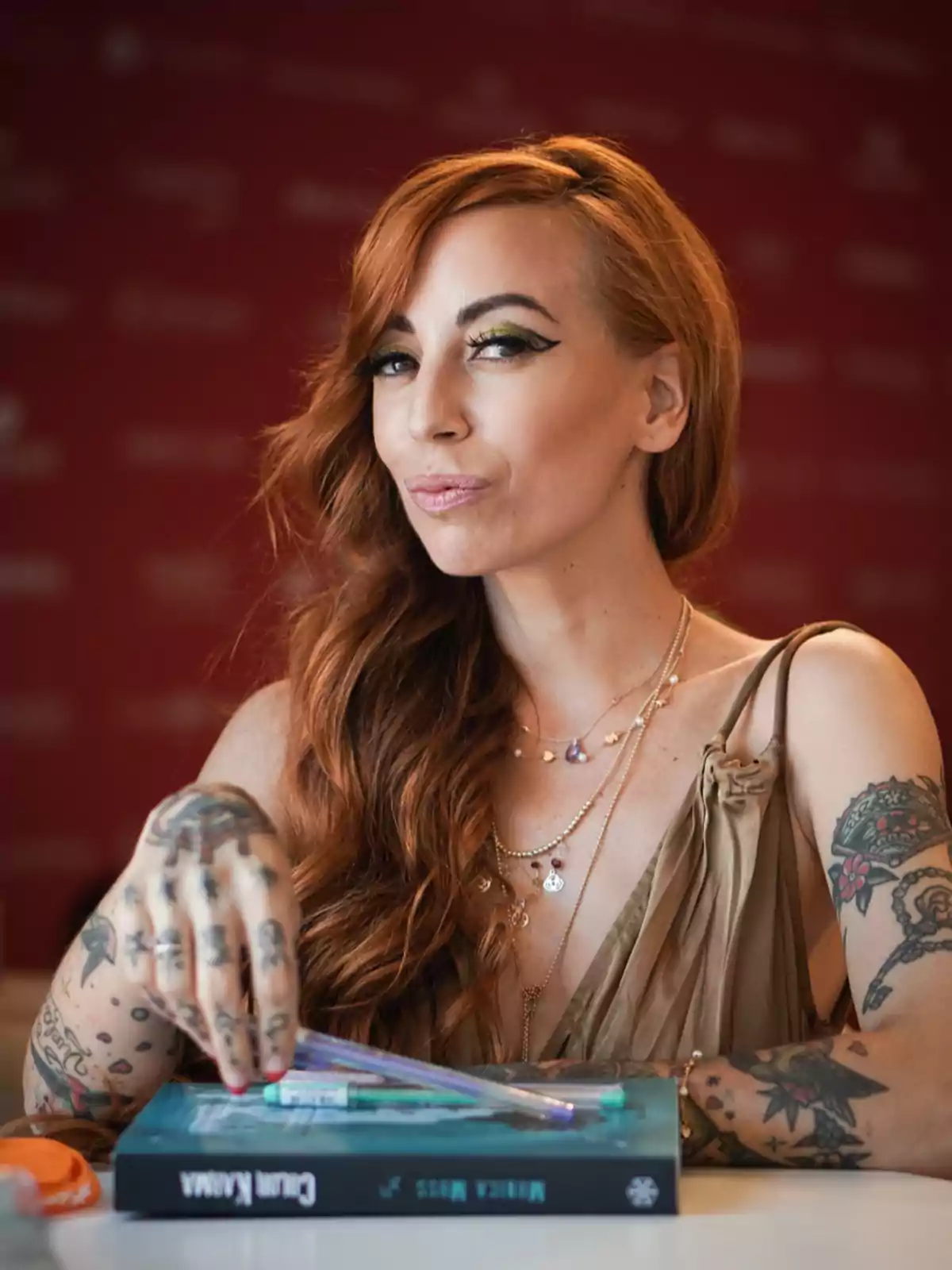 Mujer pelirroja con tatuajes y maquillaje llamativo sentada frente a un libro y bolígrafos sobre una mesa Mujer pelirroja con tatuajes y maquillaje llamativo sentada frente a un libro y bolígrafos sobre una mesa