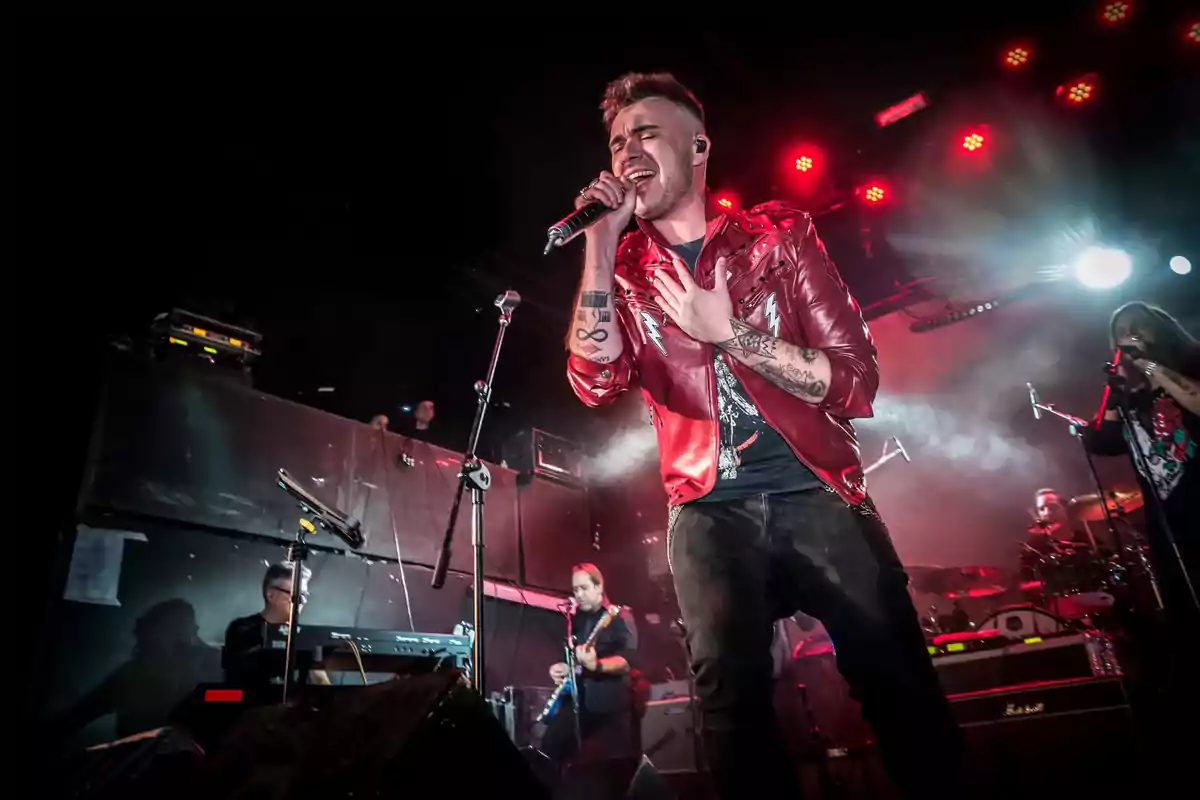 Cantante con chaqueta roja interpretando una canción en el escenario acompañado de una banda y luces rojas de fondo