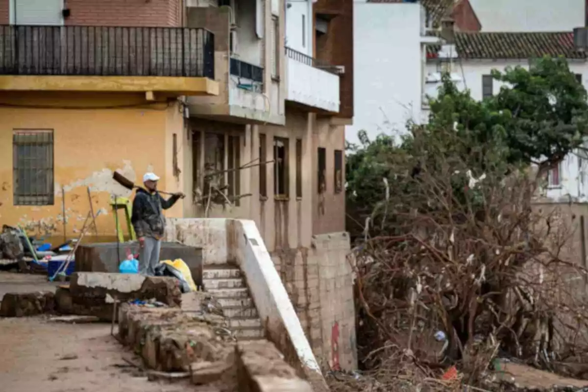 Un hombre con gorra blanca y chaqueta oscura sostiene una escoba mientras está de pie en una calle con escombros y edificios dañados a su alrededor en Paiporta.