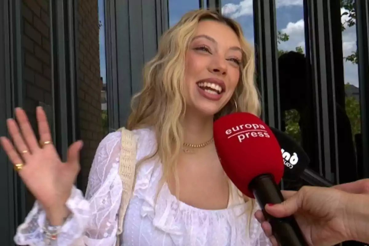 Alejandra Rubio sonriente con blusa blanca siendo entrevistada por un micrófono rojo de Europa Press mientras saluda con la mano Alejandra Rubio sonriente con blusa blanca siendo entrevistada por un micrófono rojo de Europa Press mientras saluda con la mano