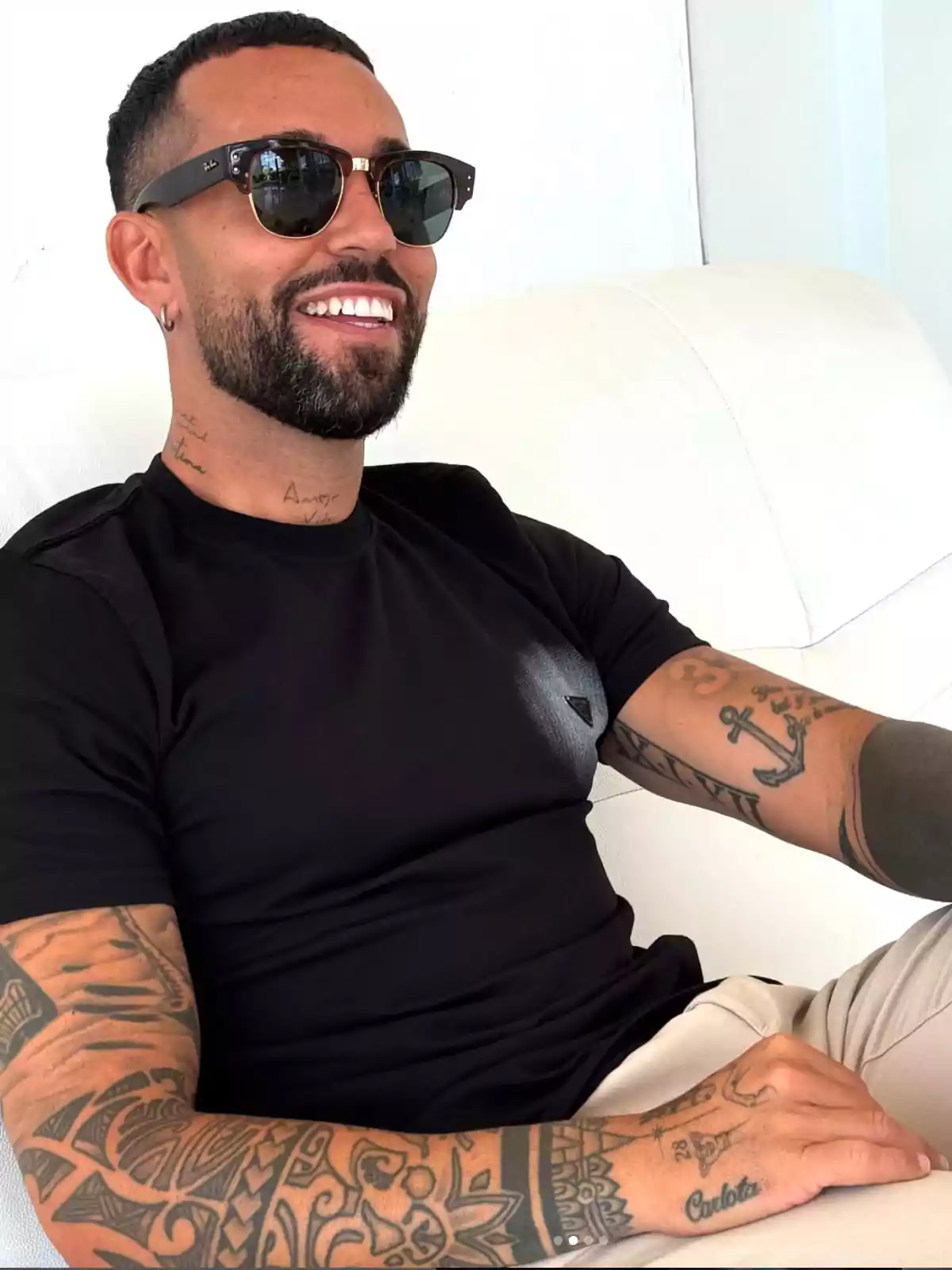 Omar Sánchez con barba y gafas de sol sentado en un sofá blanco, lleva camiseta negra y tiene varios tatuajes en los brazos y el cuello Omar Sánchez con barba y gafas de sol sentado en un sofá blanco, lleva camiseta negra y tiene varios tatuajes en los brazos y el cuello