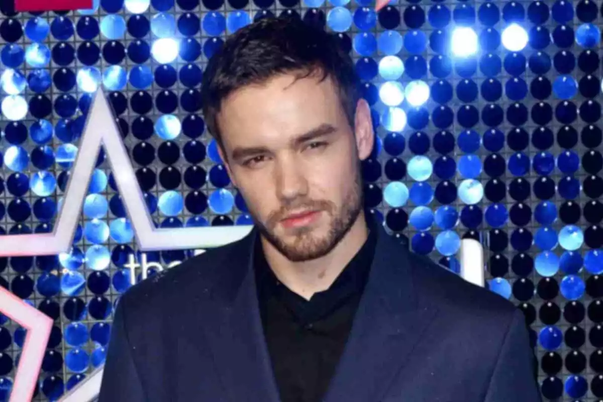 Liam Payne con barba y cabello corto lleva un traje oscuro frente a un fondo de luces azules.