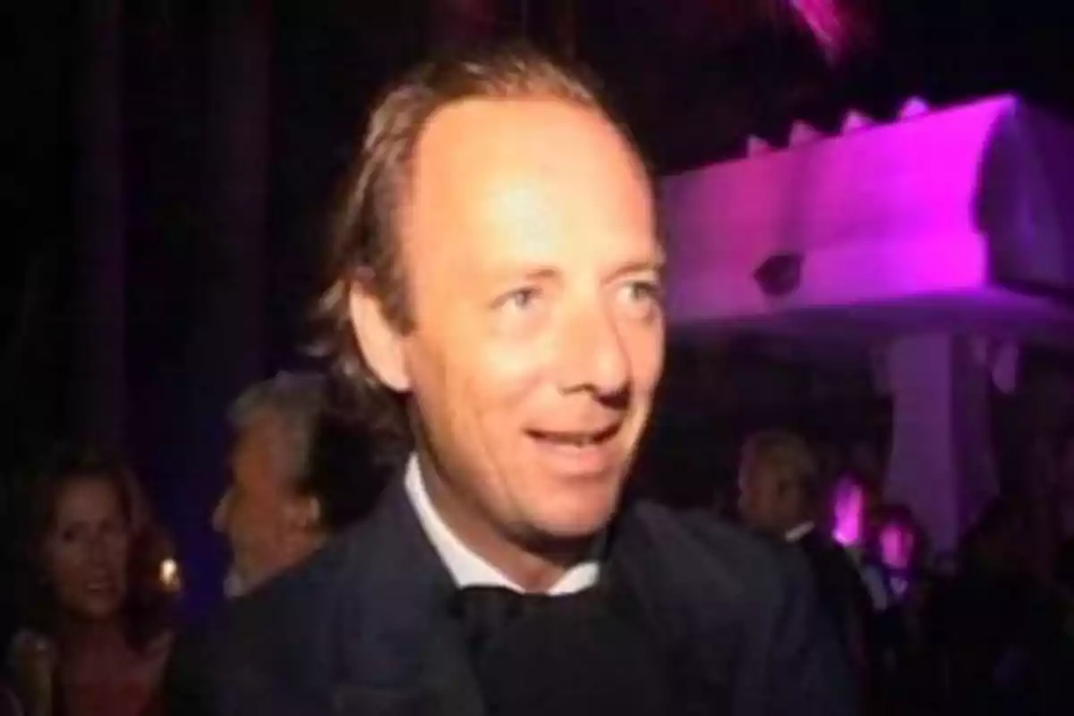 Hombre de cabello claro y corto vestido de traje oscuro en un evento nocturno con luces moradas y personas al fondo Hombre de cabello claro y corto vestido de traje oscuro en un evento nocturno con luces moradas y personas al fondo