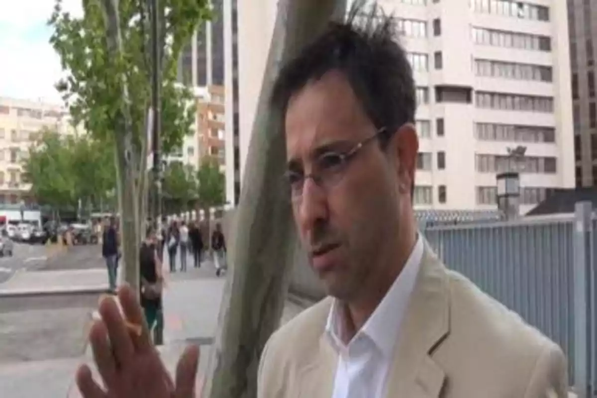 Hombre con gafas y chaqueta clara hablando al aire libre en una ciudad con edificios y árboles al fondo Hombre con gafas y chaqueta clara hablando al aire libre en una ciudad con edificios y árboles al fondo