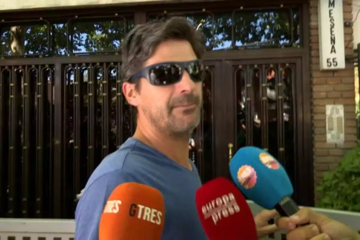 Un hombre con gafas de sol y barba es entrevistado por varios medios de comunicación frente a una puerta de metal.