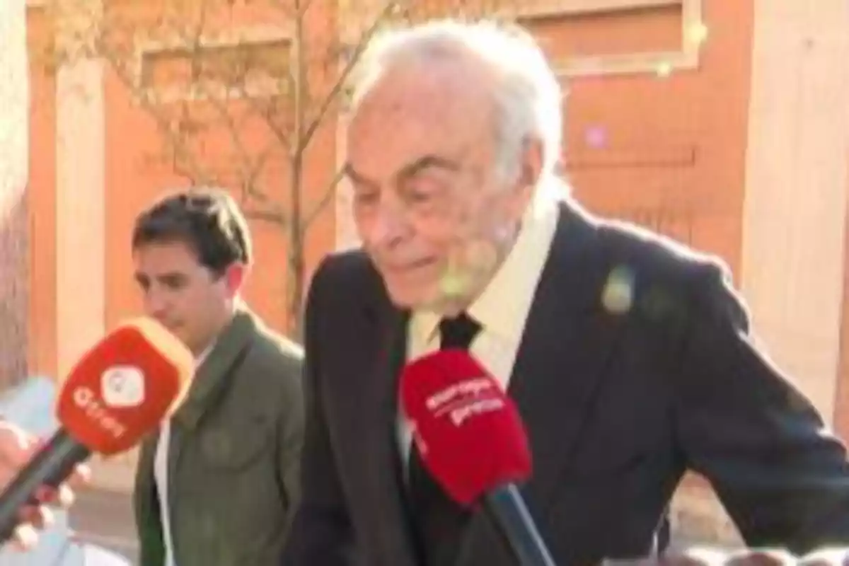 Un hombre mayor con traje oscuro es entrevistado por periodistas en la calle mientras otra persona camina detrás de él Un hombre mayor con traje oscuro es entrevistado por periodistas en la calle mientras otra persona camina detrás de él