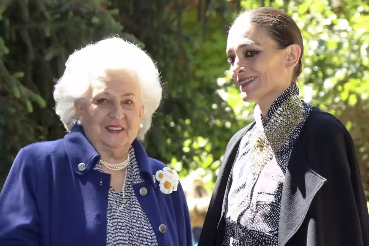 Dos mujeres posan juntas al aire libre, una de ellas con cabello blanco y chaqueta azul, la otra con cabello recogido y atuendo oscuro. Dos mujeres posan juntas al aire libre, una de ellas con cabello blanco y chaqueta azul, la otra con cabello recogido y atuendo oscuro.