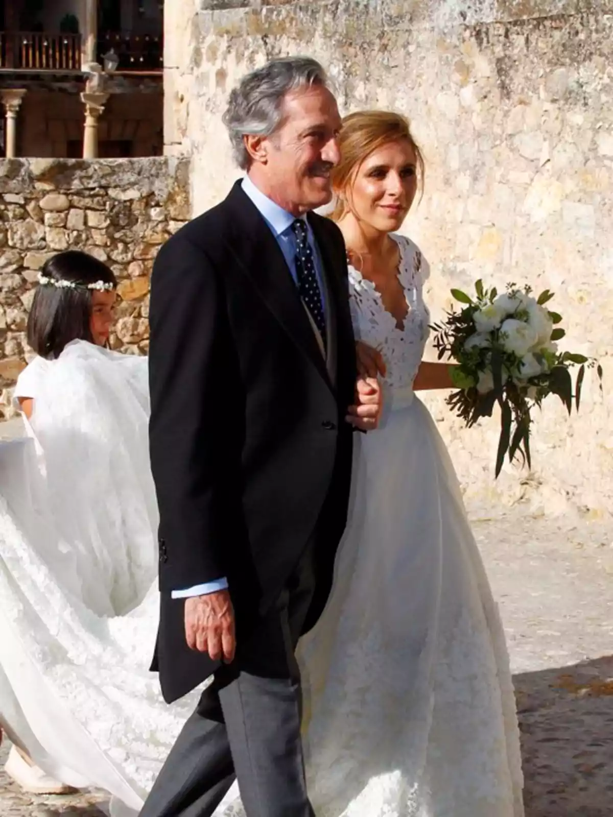 Una novia con vestido blanco camina del brazo de un hombre mayor vestido de traje oscuro mientras una niña sostiene la cola del vestido en un entorno de piedra. Una novia con vestido blanco camina del brazo de un hombre mayor vestido de traje oscuro mientras una niña sostiene la cola del vestido en un entorno de piedra.