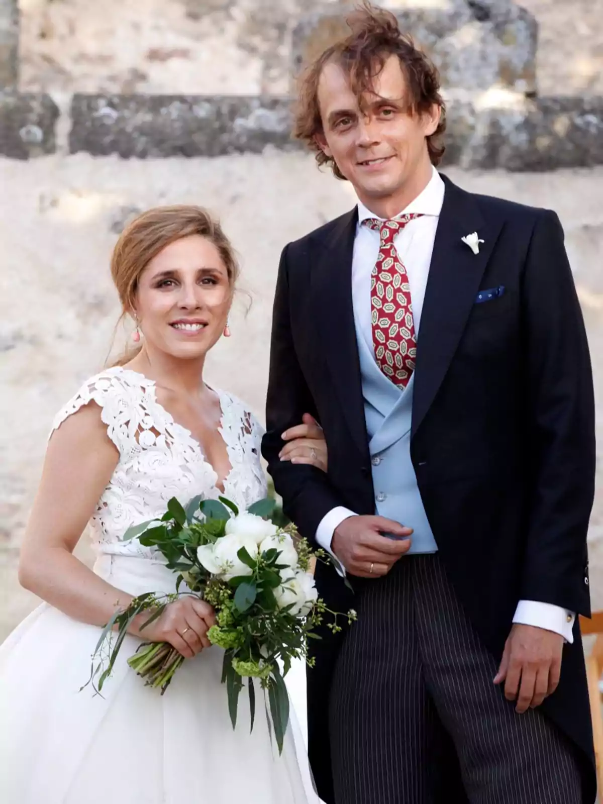 Una pareja vestida de novios sonríe mientras posa para una foto; la novia lleva un vestido blanco con encaje y sostiene un ramo de flores, mientras que el novio viste un traje formal con chaleco azul y corbata estampada. Una pareja vestida de novios sonríe mientras posa para una foto; la novia lleva un vestido blanco con encaje y sostiene un ramo de flores, mientras que el novio viste un traje formal con chaleco azul y corbata estampada.