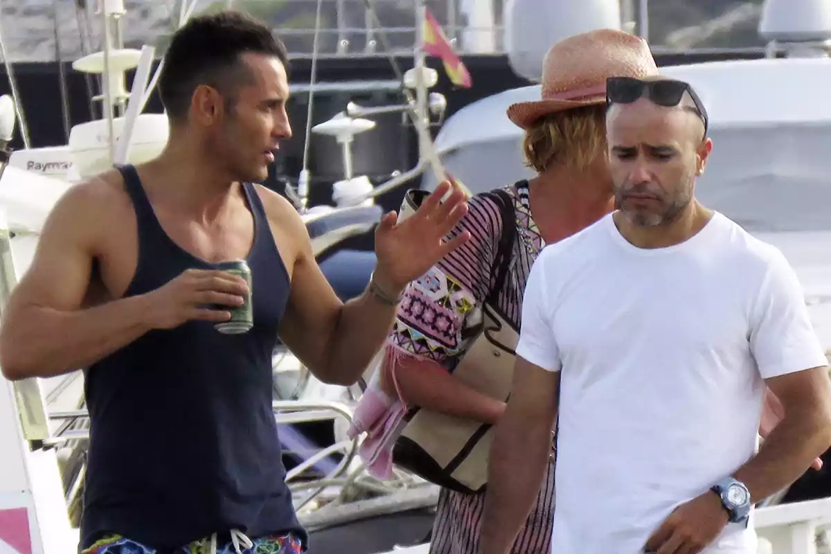 Tres personas caminan por un muelle con yates al fondo, una de ellas lleva una lata en la mano y otra viste una camiseta blanca y gafas de sol en la cabeza Tres personas caminan por un muelle con yates al fondo, una de ellas lleva una lata en la mano y otra viste una camiseta blanca y gafas de sol en la cabeza