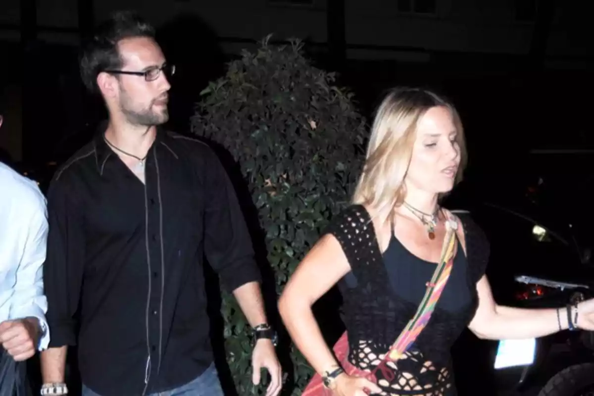 Dos personas caminando de noche por la calle, una mujer rubia al frente y un hombre con gafas detrás, ambos vestidos de manera casual Dos personas caminando de noche por la calle, una mujer rubia al frente y un hombre con gafas detrás, ambos vestidos de manera casual