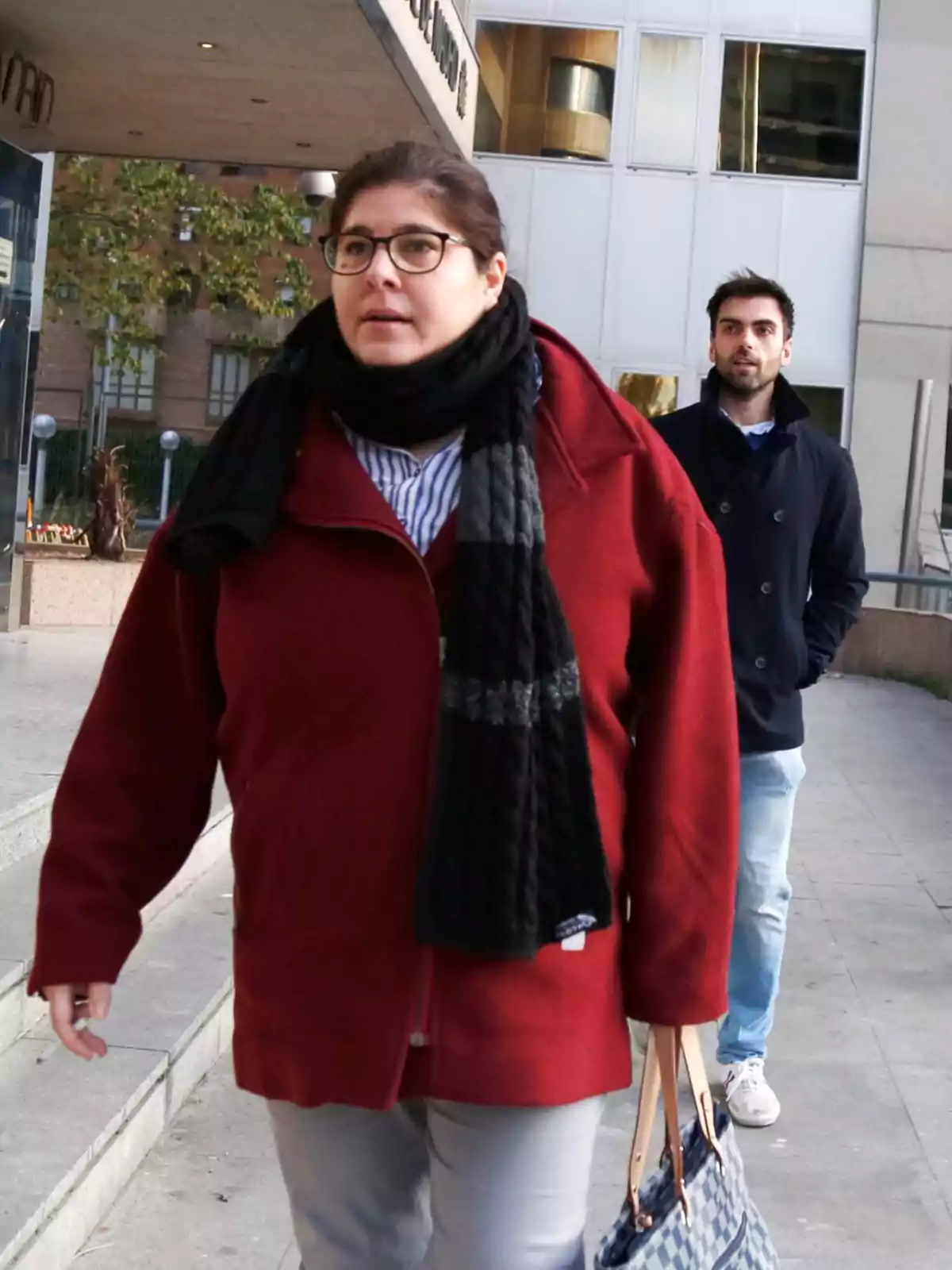 Mujer con abrigo rojo y bufanda negra caminando por la acera frente a un edificio, seguida por un hombre con chaqueta oscura y pantalón claro Mujer con abrigo rojo y bufanda negra caminando por la acera frente a un edificio, seguida por un hombre con chaqueta oscura y pantalón claro