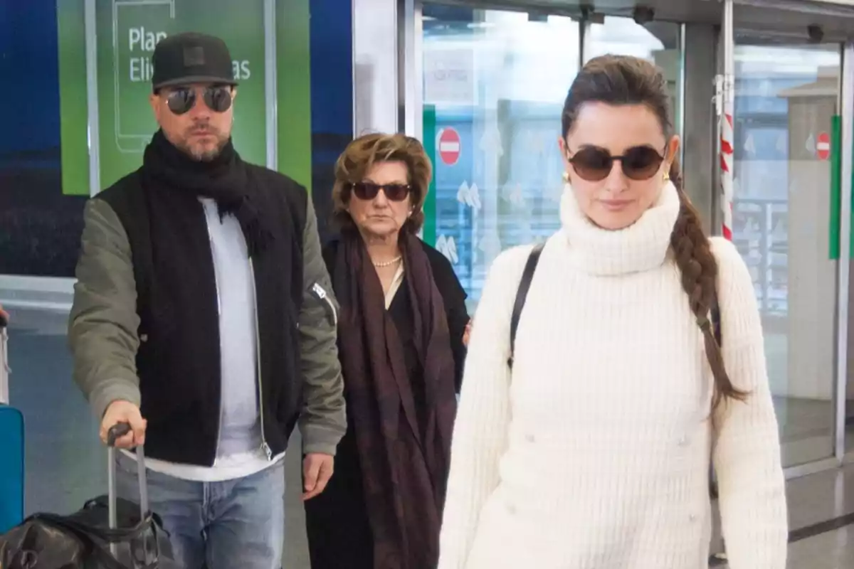 Tres personas con gafas de sol caminan en un aeropuerto, una mujer lleva un suéter blanco de cuello alto y el hombre lleva una chaqueta oscura y gorra. Tres personas con gafas de sol caminan en un aeropuerto, una mujer lleva un suéter blanco de cuello alto y el hombre lleva una chaqueta oscura y gorra.