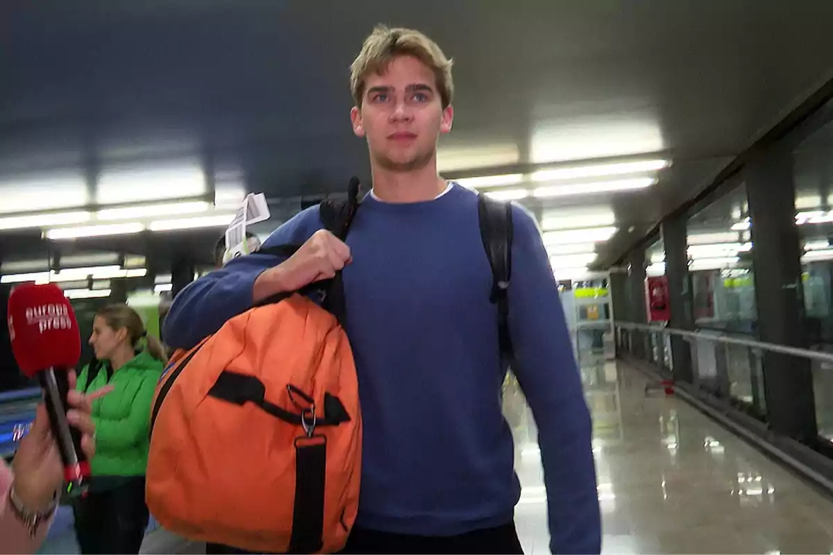 Un hombre joven con suéter azul y mochila naranja camina por un pasillo iluminado mientras una persona sostiene un micrófono cerca. Un hombre joven con suéter azul y mochila naranja camina por un pasillo iluminado mientras una persona sostiene un micrófono cerca.