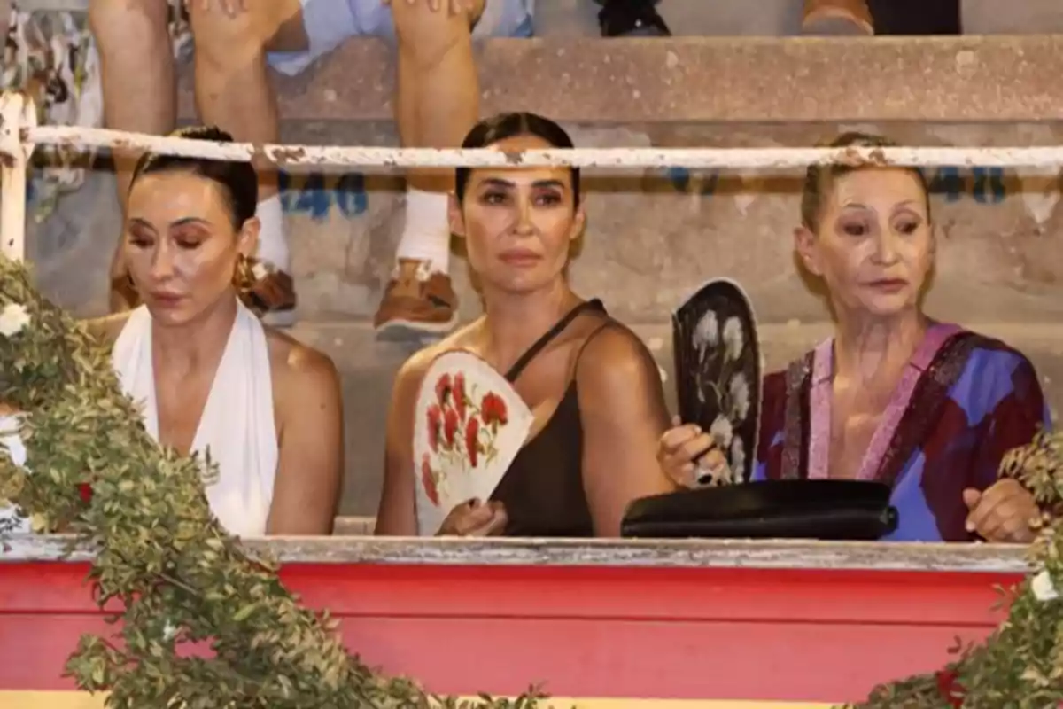 Tres mujeres sentadas en una plaza de toros sosteniendo abanicos mientras observan el evento. Tres mujeres sentadas en una plaza de toros sosteniendo abanicos mientras observan el evento.