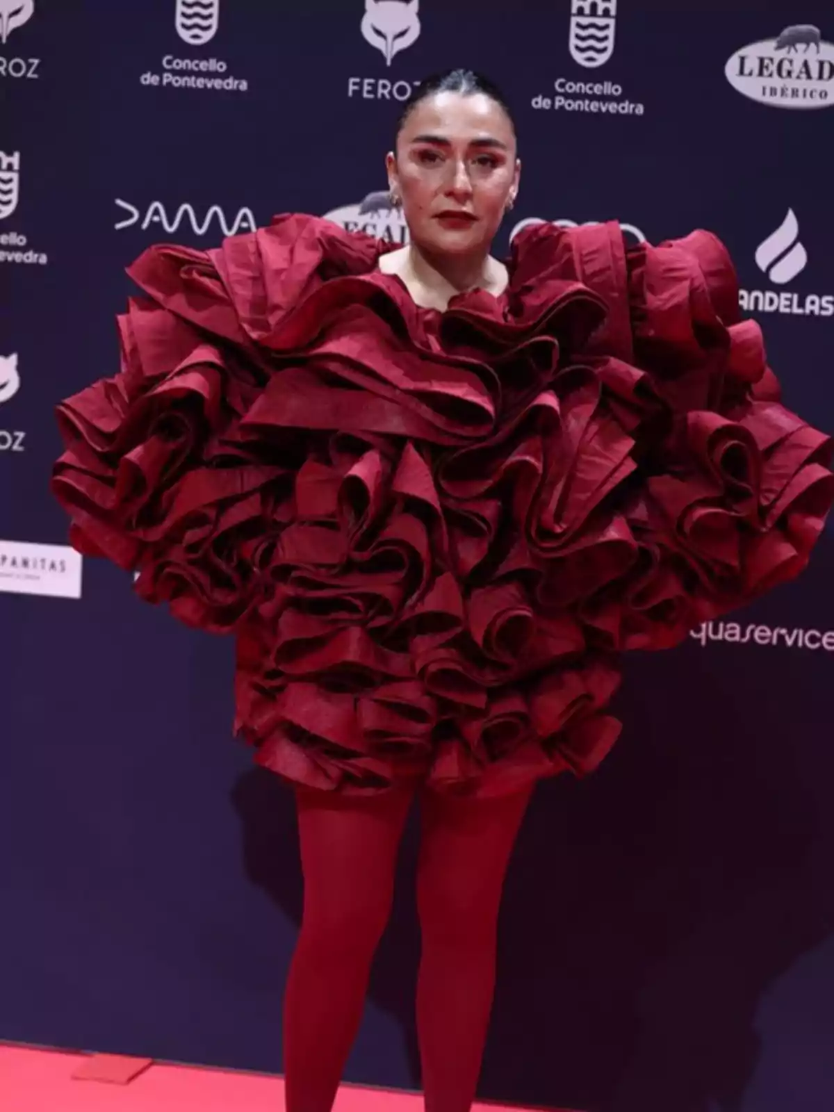 Una persona con un vestido rojo voluminoso y medias del mismo color posa en una alfombra roja frente a un fondo con logotipos de patrocinadores.