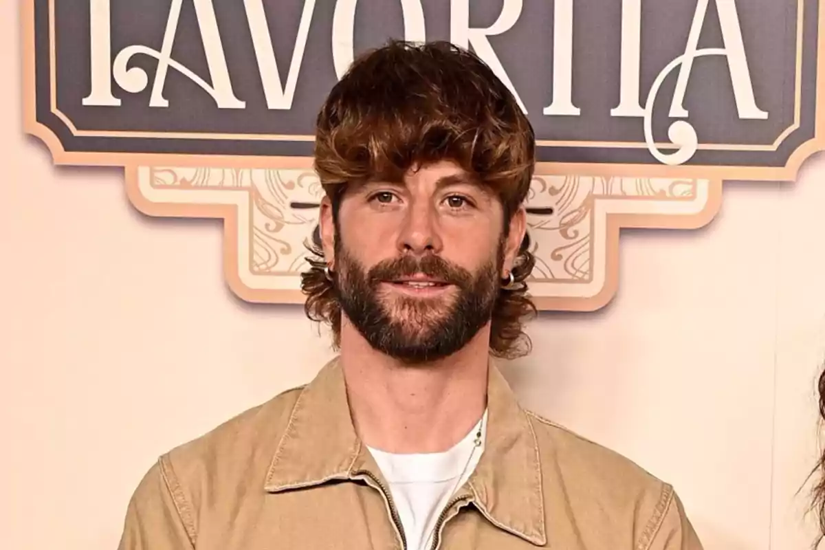 Un hombre con barba y cabello rizado lleva una chaqueta beige frente a un cartel decorativo.