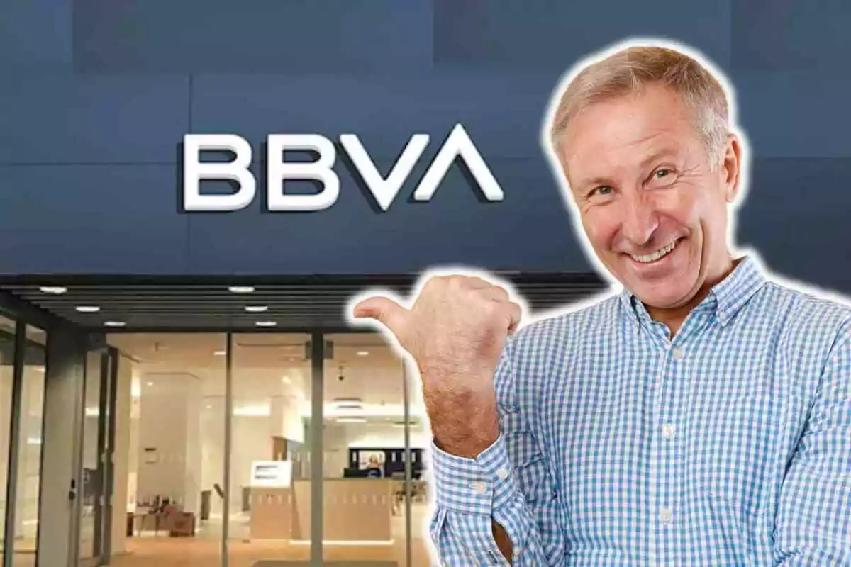 Un hombre sonriente con camisa a cuadros azules y blancos señala con el pulgar hacia una sucursal del banco BBVA.