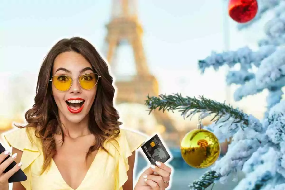 Mujer sonriente con gafas amarillas sosteniendo una tarjeta de crédito y un teléfono frente a un árbol de Navidad nevado y la Torre Eiffel de fondo.