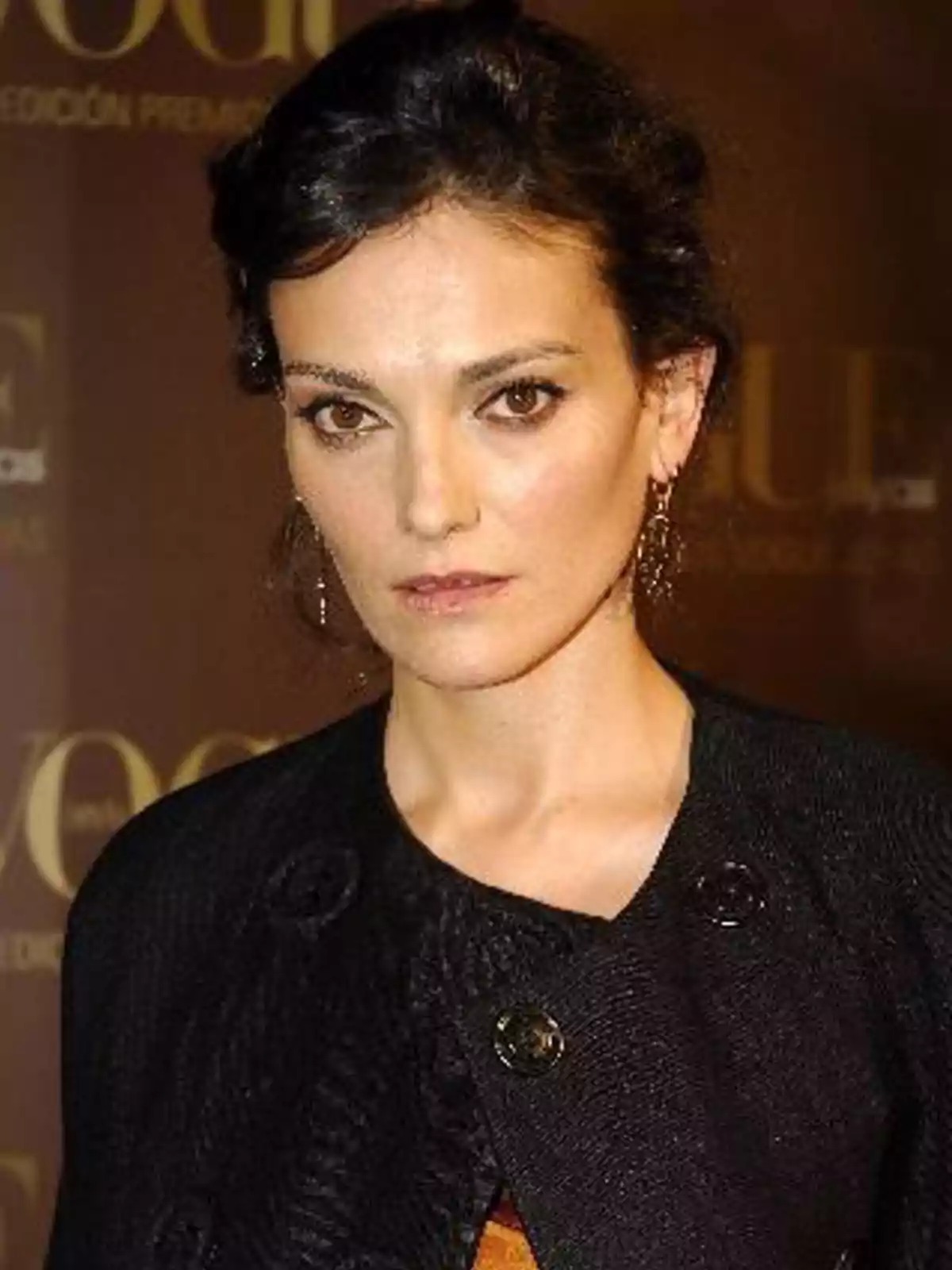 Mujer de cabello oscuro y recogido con chaqueta negra posando frente a un fondo marrón en un evento elegante Mujer de cabello oscuro y recogido con chaqueta negra posando frente a un fondo marrón en un evento elegante