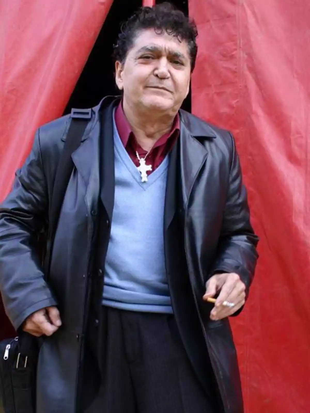 Un hombre de pie frente a una cortina roja, vistiendo un abrigo de cuero negro, suéter azul y camisa roja, sosteniendo un cigarrillo en una mano y una bolsa en la otra. Un hombre de pie frente a una cortina roja, vistiendo un abrigo de cuero negro, suéter azul y camisa roja, sosteniendo un cigarrillo en una mano y una bolsa en la otra.