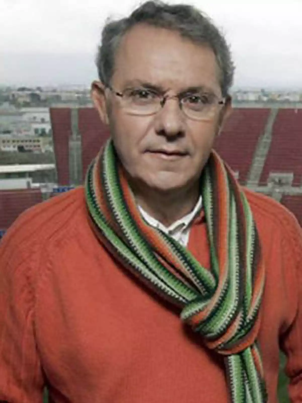 Un hombre con gafas y cabello canoso lleva un suéter naranja y una bufanda de rayas verdes y naranjas, con un estadio de fondo. Un hombre con gafas y cabello canoso lleva un suéter naranja y una bufanda de rayas verdes y naranjas, con un estadio de fondo.