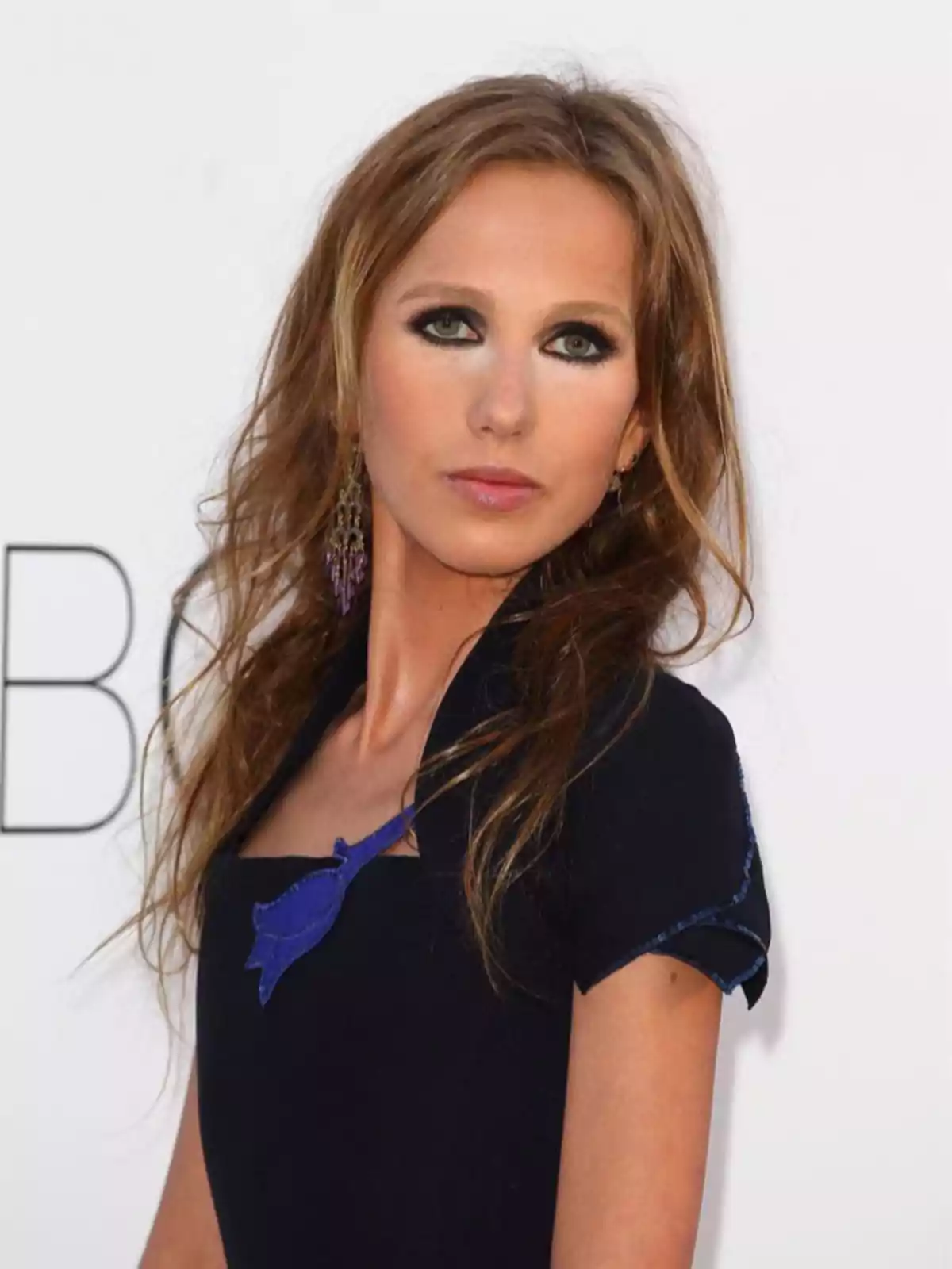 Una mujer con cabello castaño y ojos maquillados, vistiendo un vestido negro con un detalle azul, posa frente a un fondo blanco. Una mujer con cabello castaño y ojos maquillados, vistiendo un vestido negro con un detalle azul, posa frente a un fondo blanco.