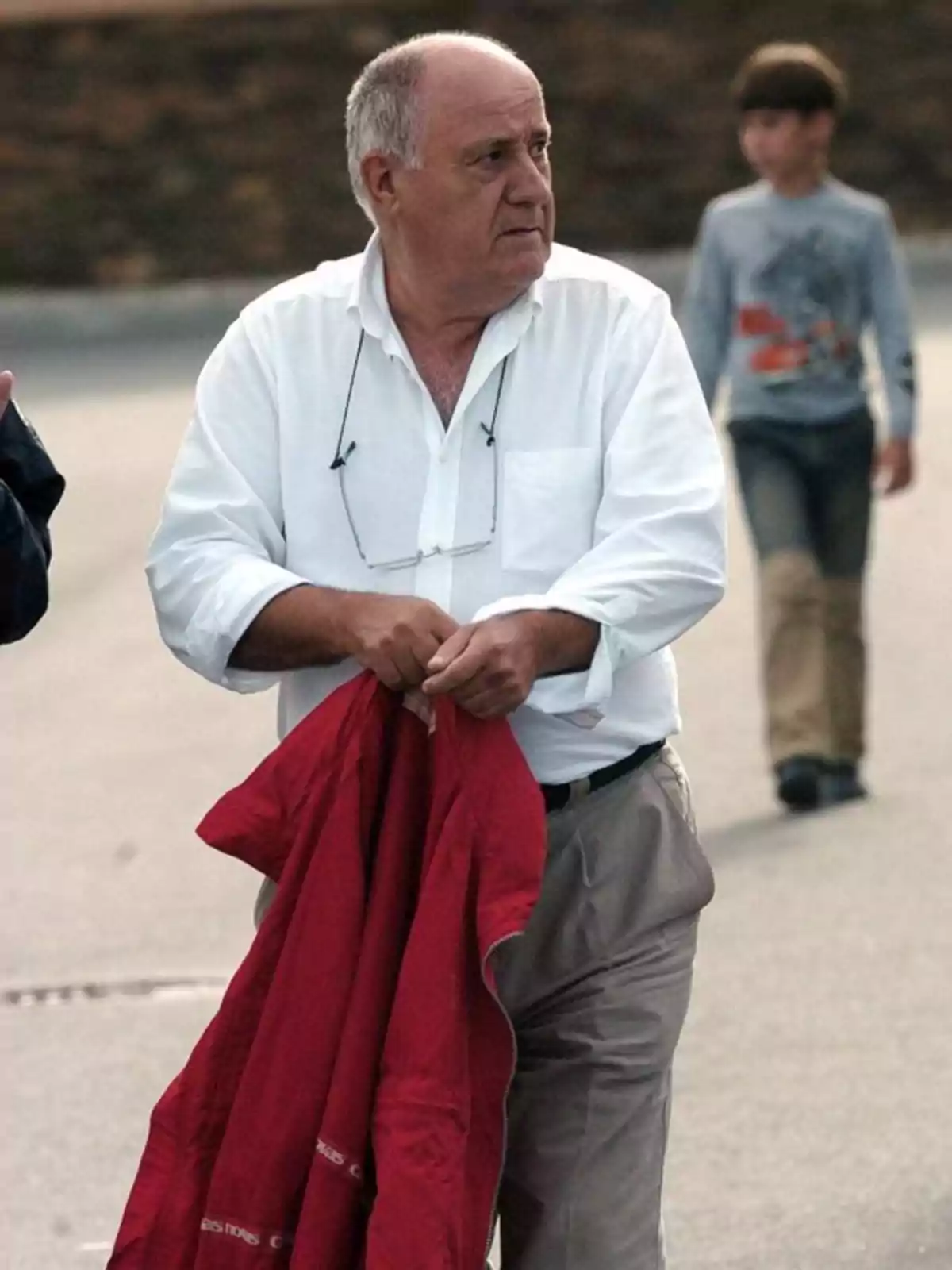 Hombre mayor de cabello canoso viste camisa blanca y pantalón beige mientras sostiene una chaqueta roja en la calle con un niño desenfocado al fondo Hombre mayor de cabello canoso viste camisa blanca y pantalón beige mientras sostiene una chaqueta roja en la calle con un niño desenfocado al fondo