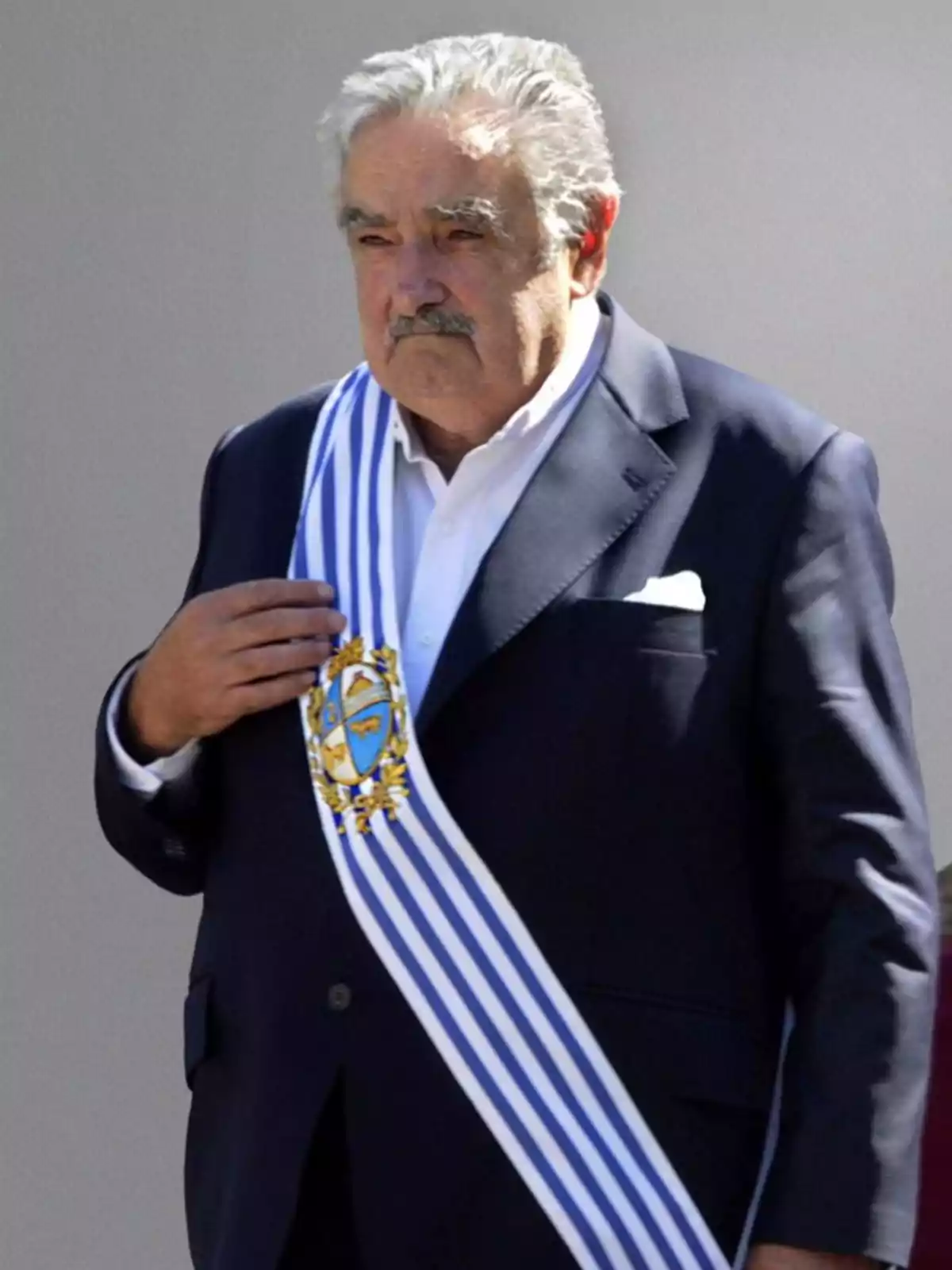 Un hombre mayor con bigote lleva un traje oscuro y una banda presidencial con rayas azules y blancas. Un hombre mayor con bigote lleva un traje oscuro y una banda presidencial con rayas azules y blancas.