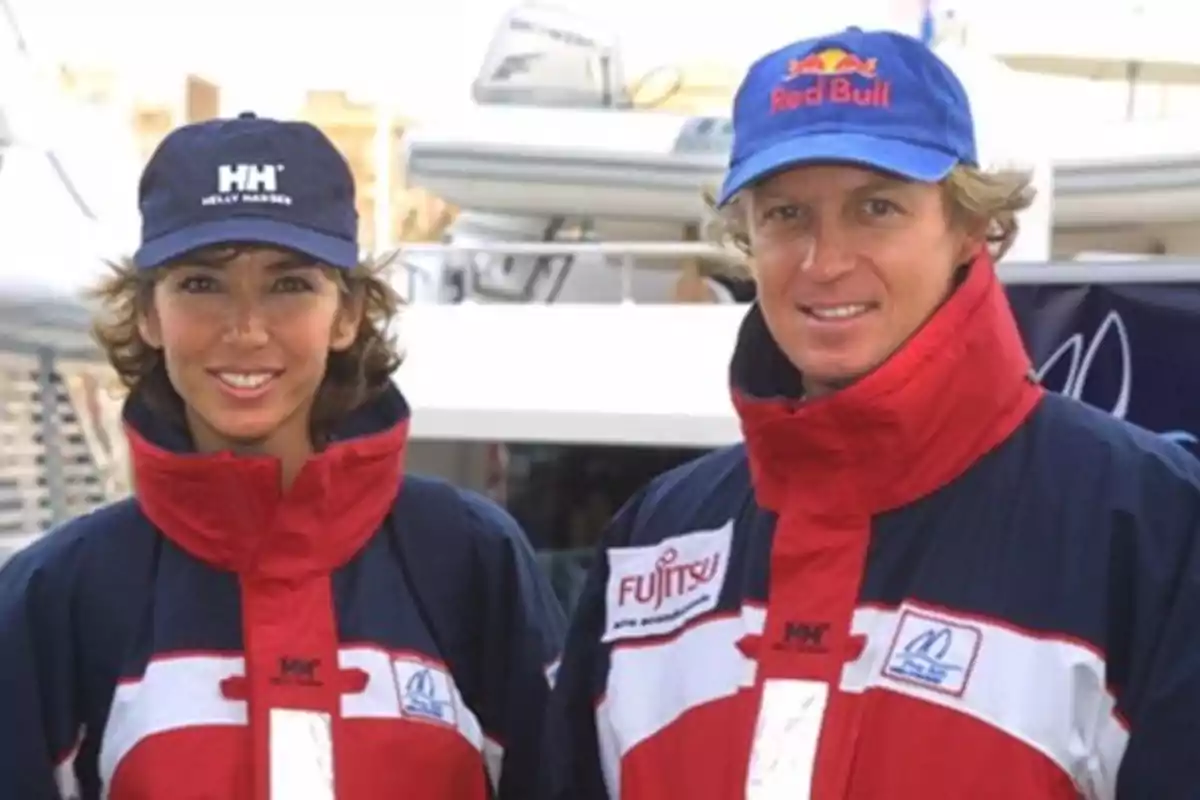 Dos personas con chaquetas y gorras deportivas posan frente a un barco Dos personas con chaquetas y gorras deportivas posan frente a un barco