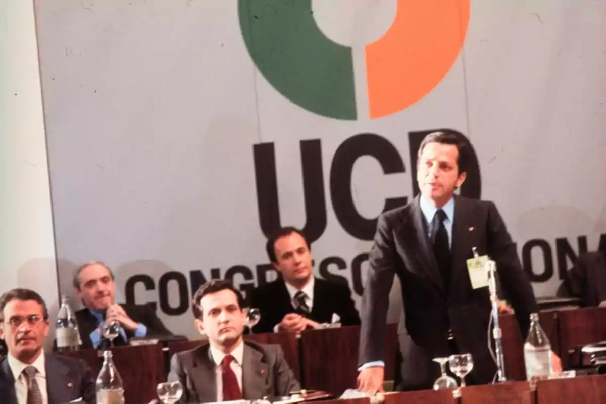 Un hombre de traje habla de pie en un congreso político con el logo de la UCD de fondo y varias personas sentadas a su alrededor Un hombre de traje habla de pie en un congreso político con el logo de la UCD de fondo y varias personas sentadas a su alrededor