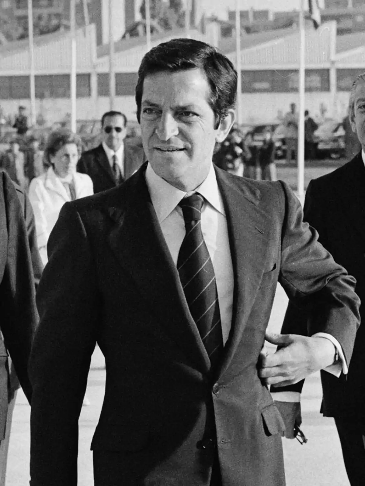 Hombre de traje oscuro y corbata caminando al aire libre con personas al fondo en una imagen en blanco y negro Hombre de traje oscuro y corbata caminando al aire libre con personas al fondo en una imagen en blanco y negro