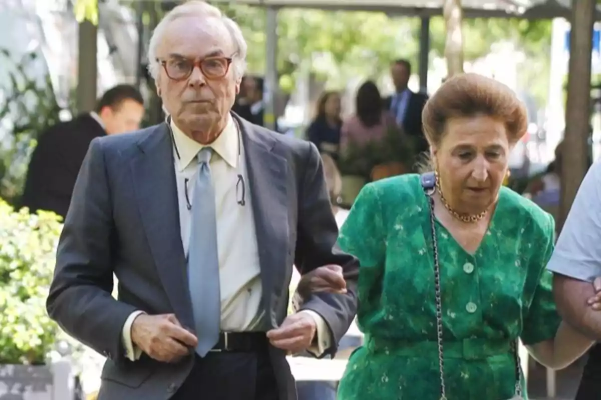 Pareja de personas mayores caminando juntas al aire libre en un entorno urbano con árboles y personas al fondo Pareja de personas mayores caminando juntas al aire libre en un entorno urbano con árboles y personas al fondo
