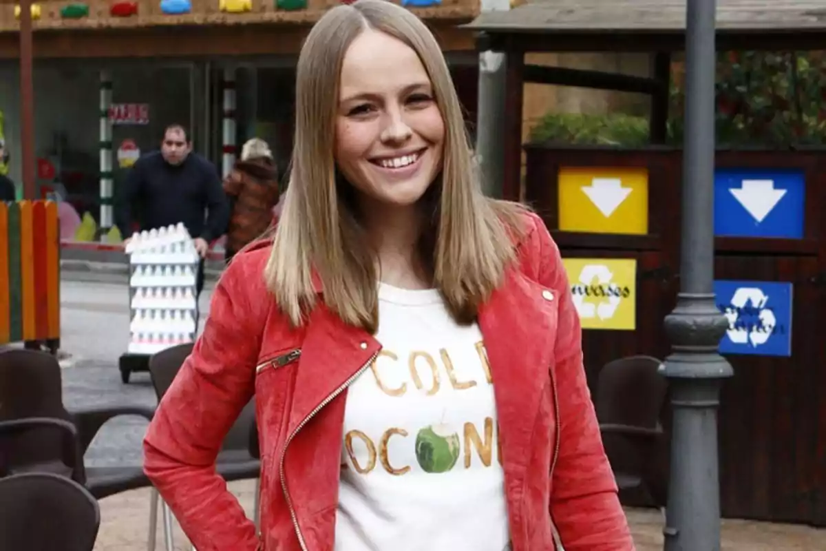 Una persona con chaqueta roja y camiseta blanca sonríe al aire libre, con un fondo de sillas y señales de reciclaje. Una persona con chaqueta roja y camiseta blanca sonríe al aire libre, con un fondo de sillas y señales de reciclaje.