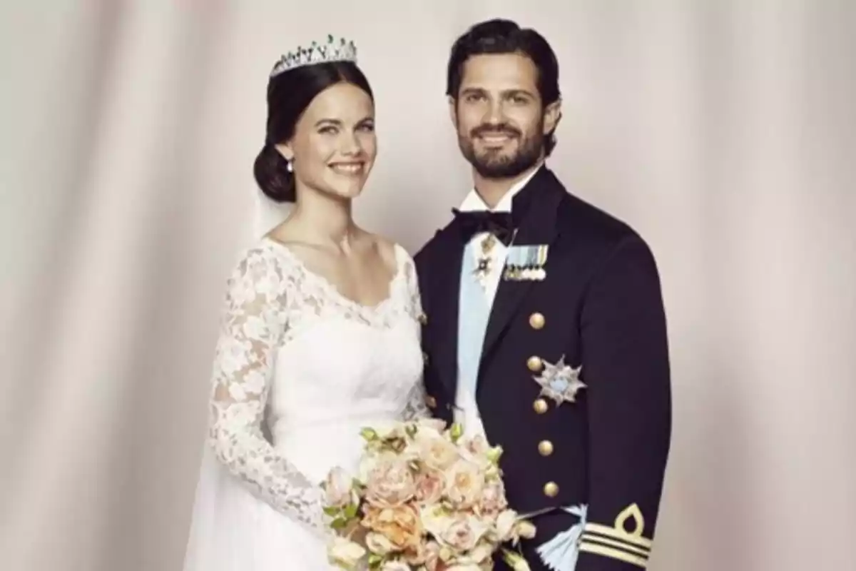 Pareja vestida de novios con la mujer usando un vestido blanco de encaje y tiara y el hombre con uniforme de gala y condecoraciones ambos sonriendo y posando juntos