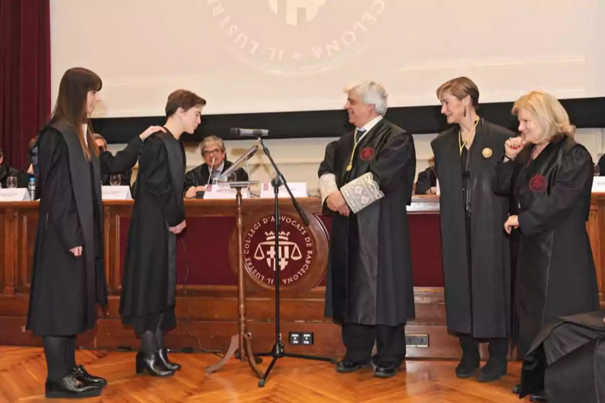 Un grupo de personas vestidas con togas negras participa en una ceremonia formal en un auditorio, donde una persona parece estar tomando un juramento frente a un podio, mientras otras observan y participan en el evento.